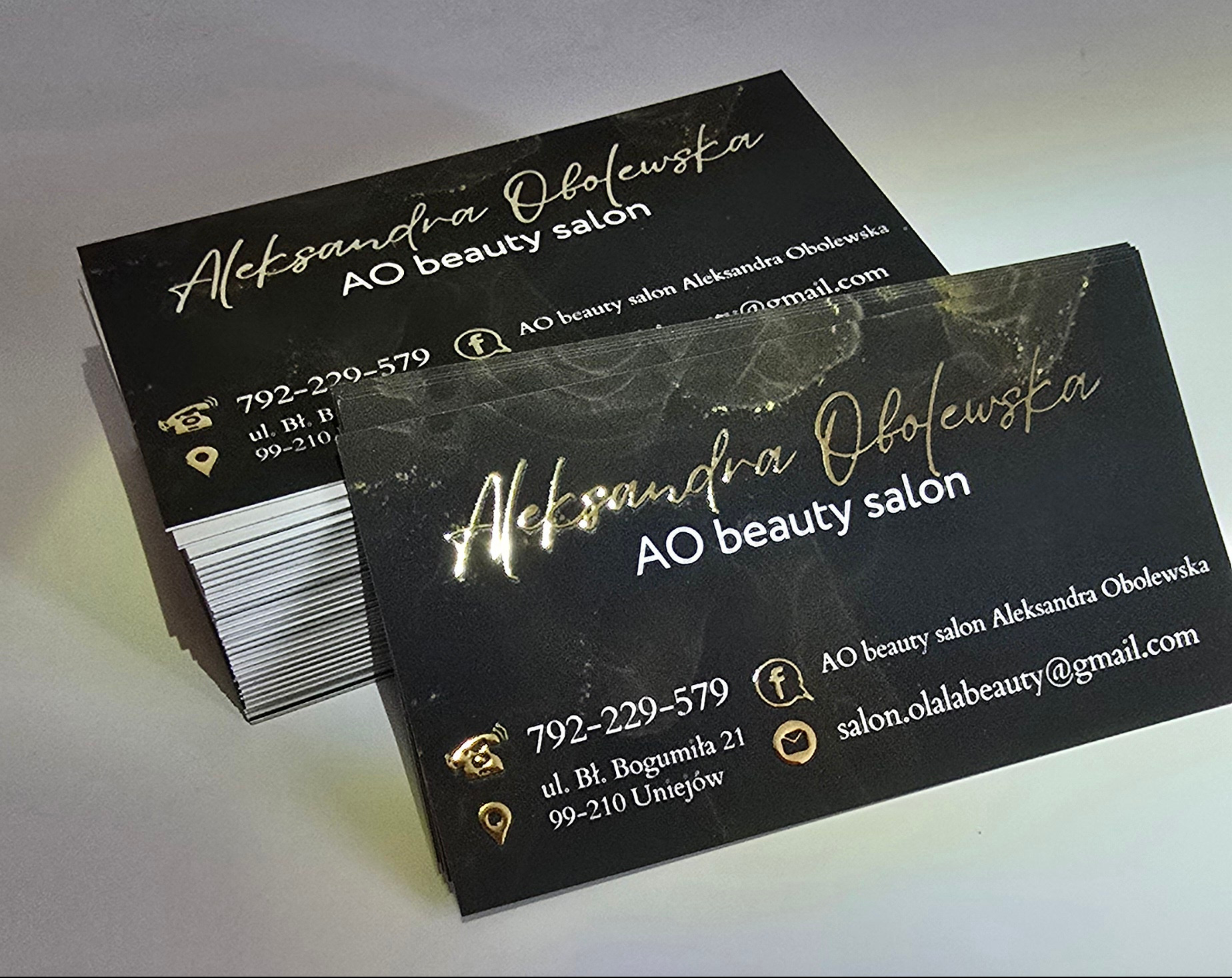 Eleganckie, czarno-złote wizytówki z połyskującym, marmurkowym tłem i danymi kontaktowymi salonu kosmetycznego, ułożone w dwie stosy na jasnym podłożu.