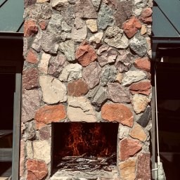 EKOLIFE.COM.PL SPÓŁKA Z OGRANICZONĄ ODPOWIEDZIALNOŚCIĄ - Kamienny kominek zewnętrzny z paleniskiem wypełnionym płomieniami, widoczny z bliska, z elementami drewnianego tarasu i zadaszenia w tle.