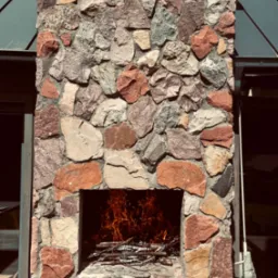 Kamienny kominek zewnętrzny z paleniskiem wypełnionym płomieniami, widoczny z bliska, z elementami drewnianego tarasu i zadaszenia w tle.