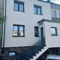 Odnowiona elewacja domu z nowymi oknami i kamienną podmur&oacute;wką, schody wejściowe z metalową barierką. Widoczny samoch&oacute;d osobowy. Antena na dachu.