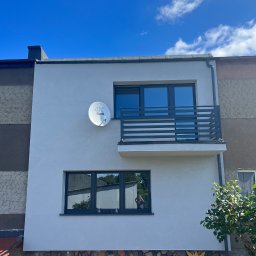 EKOLIFE.COM.PL SPÓŁKA Z OGRANICZONĄ ODPOWIEDZIALNOŚCIĄ - Dwukondygnacyjny dom z balkonem i anteną satelitarną na elewacji, widok z dołu na tle błękitnego nieba z chmurami. Część budynku wykończona kamieniem.