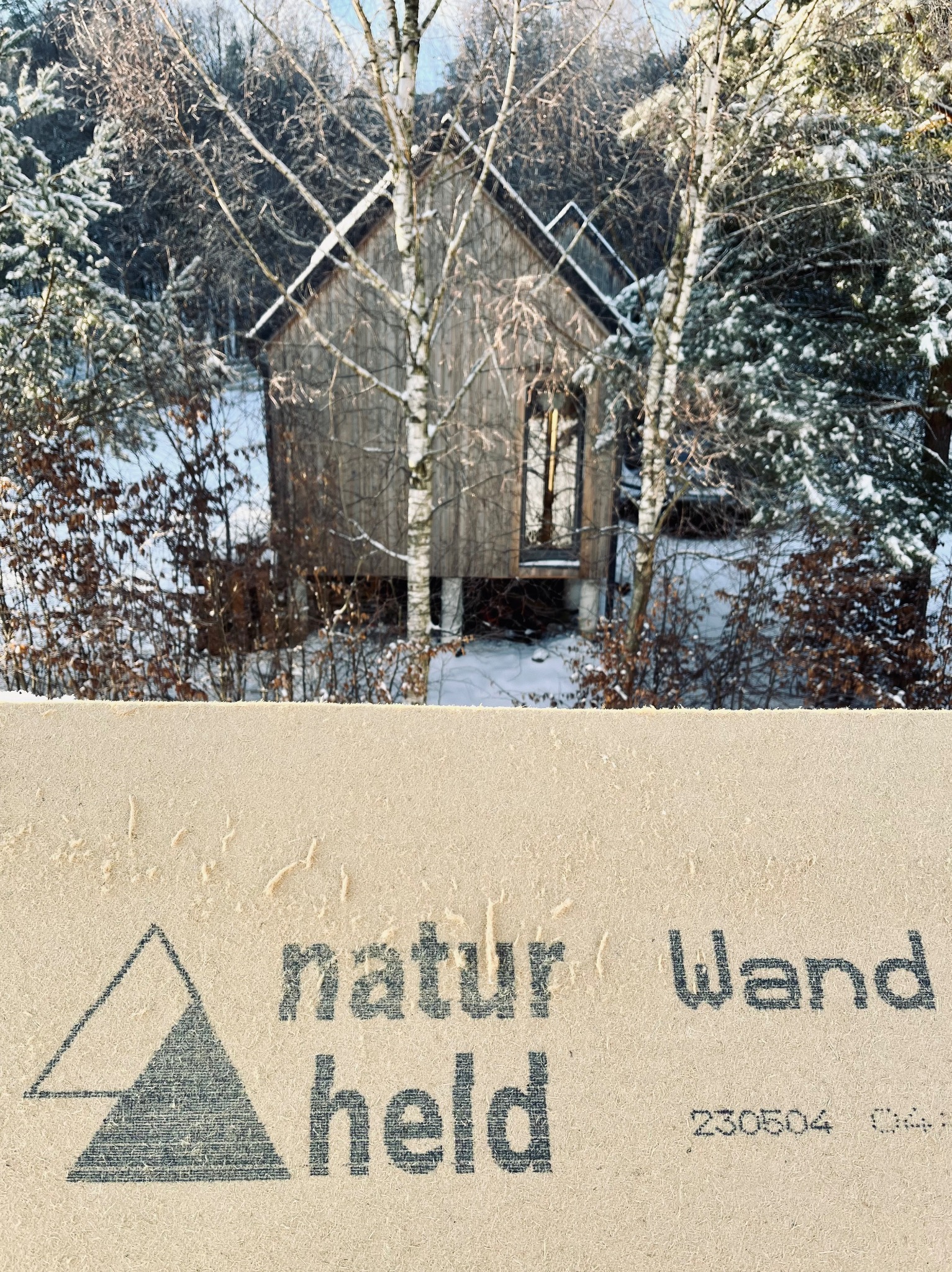 Mały, drewniany domek na palach w zimowym lesie, widziany zza próbki materiału budowlanego z nadrukiem 'natur Wand held'.