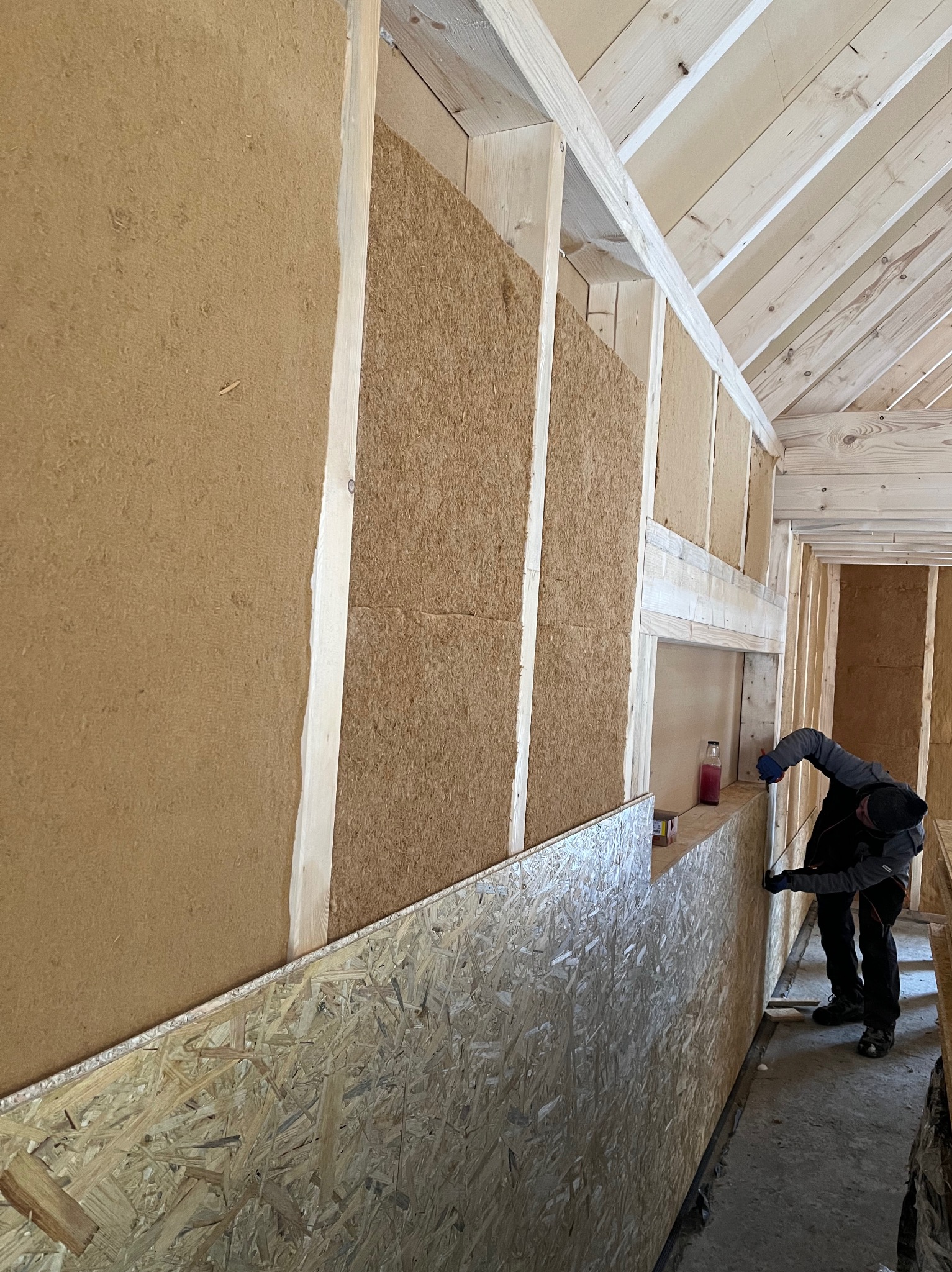 Prace wykończeniowe wewnątrz domu szkieletowego: montaż izolacji termicznej ścian przez pracownika w ubraniu roboczym. Widoczne płyty OSB i drewniana konstrukcja.