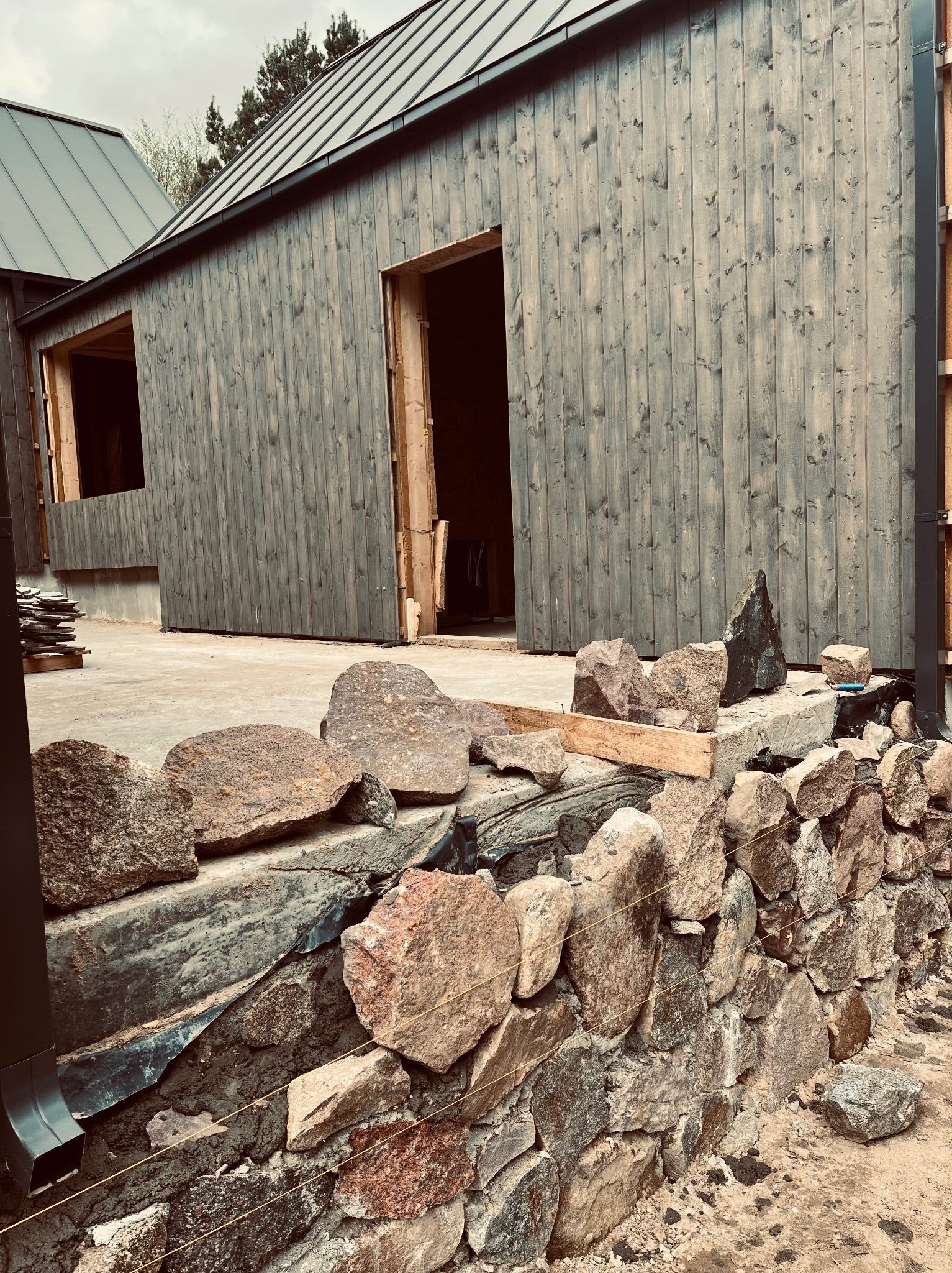 Budowa domu szkieletowego z elewacją z desek w odcieniach szarości i kamiennym murem w trakcie wznoszenia. Widoczne surowe materiały i elementy konstrukcyjne.