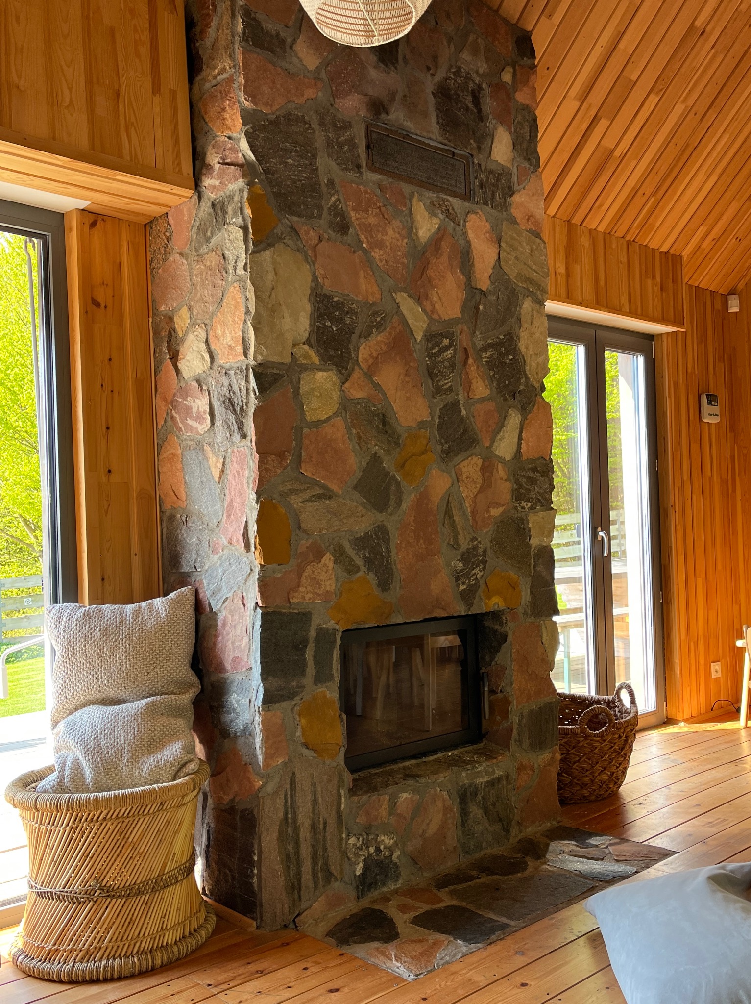 Kamienny kominek w drewnianym domu szkieletowym z widokiem na zewnątrz przez okno. Wnętrze z naturalnych materiałów.