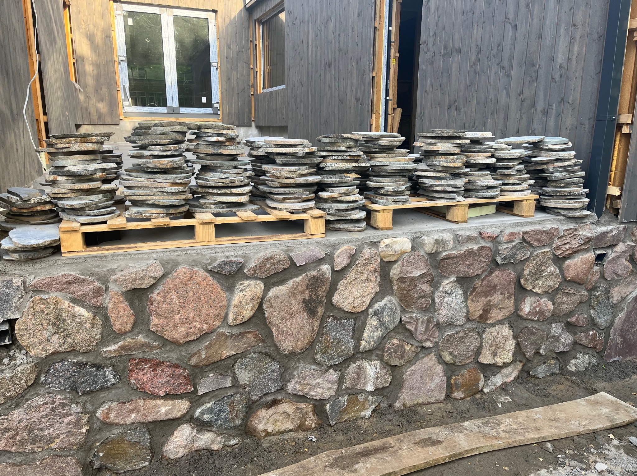 Stosy kamiennych płyt na palecie przed budynkiem z szarą drewnianą elewacją i kamienną podmurówką. Przygotowanie do prac wykończeniowych na zewnątrz.