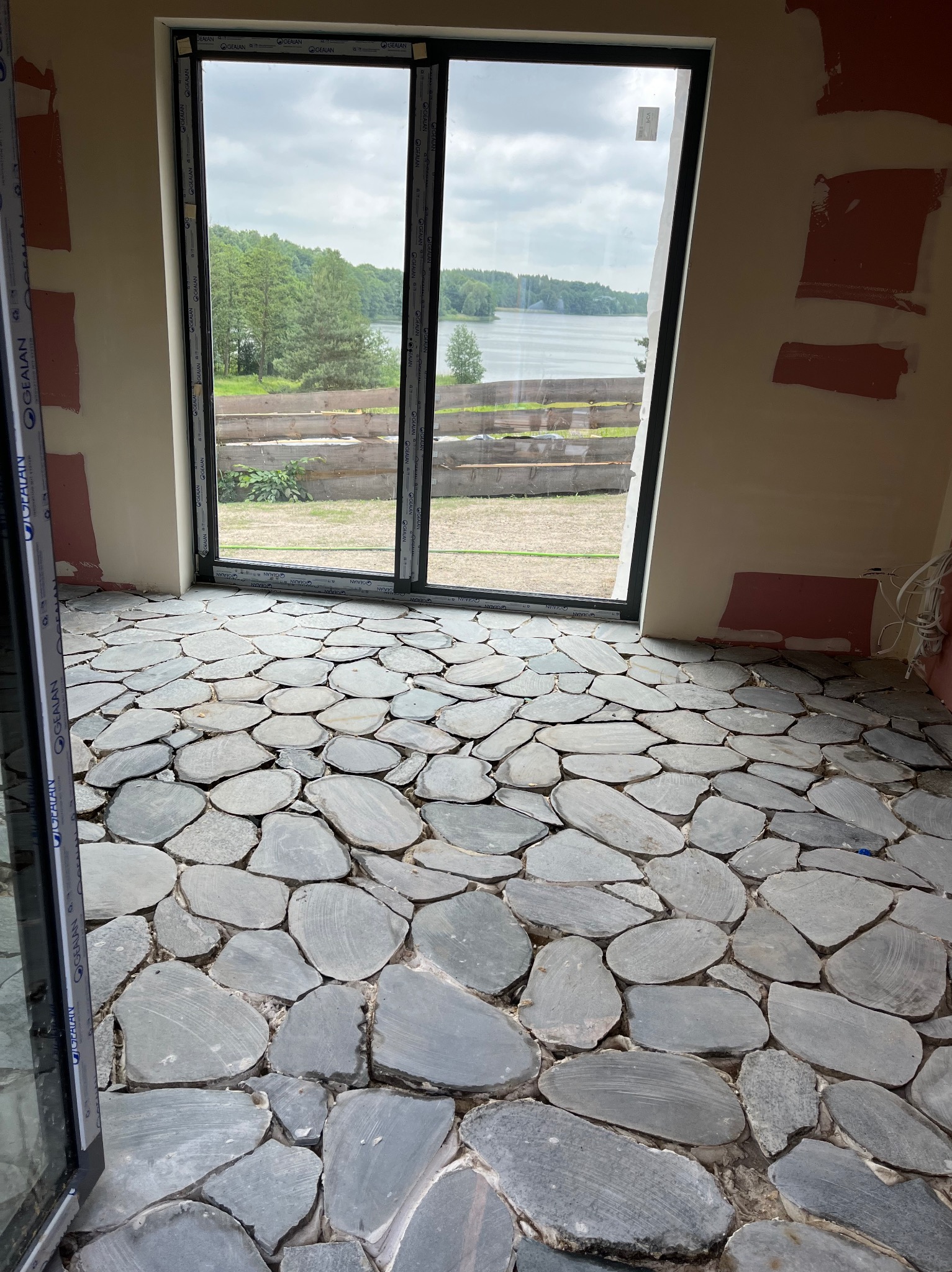 Wnętrze w trakcie wykańczania z podłogą z nieregularnych kamieni. Widok na jezioro przez duże okno przesuwne. Fragmenty ścian wymagają wykończenia.