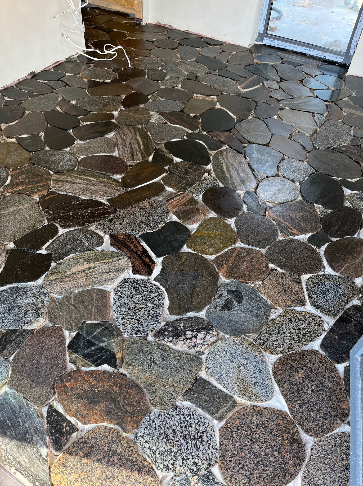 Kamienna posadzka z nieregularnych, różnokolorowych otoczaków, wypełniona jasną fugą. Widoczne fragmenty ścian i okna. Unikalny wzór kamieni.