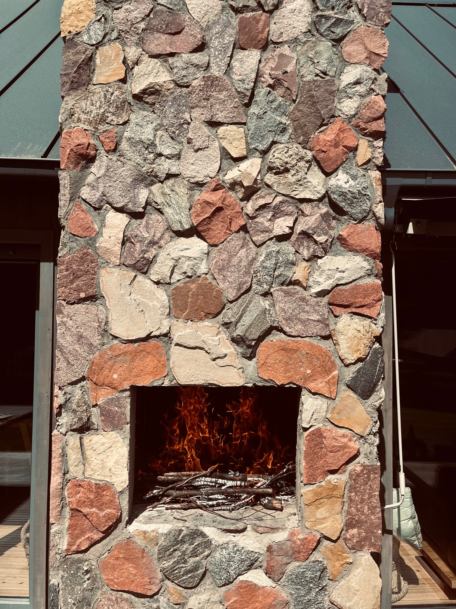 Kamienny kominek zewnętrzny z paleniskiem wypełnionym płomieniami, widoczny z bliska, z elementami drewnianego tarasu i zadaszenia w tle.