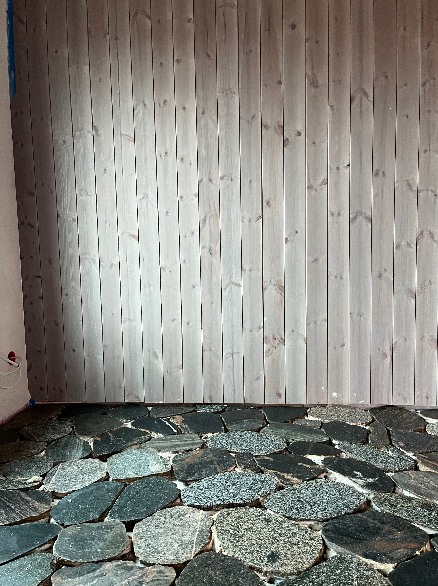 Wnętrze z wyłożoną kamienną podłogą i ścianą z paneli drewnianych w pionowym układzie. Widoczne niedokończone prace wykończeniowe.