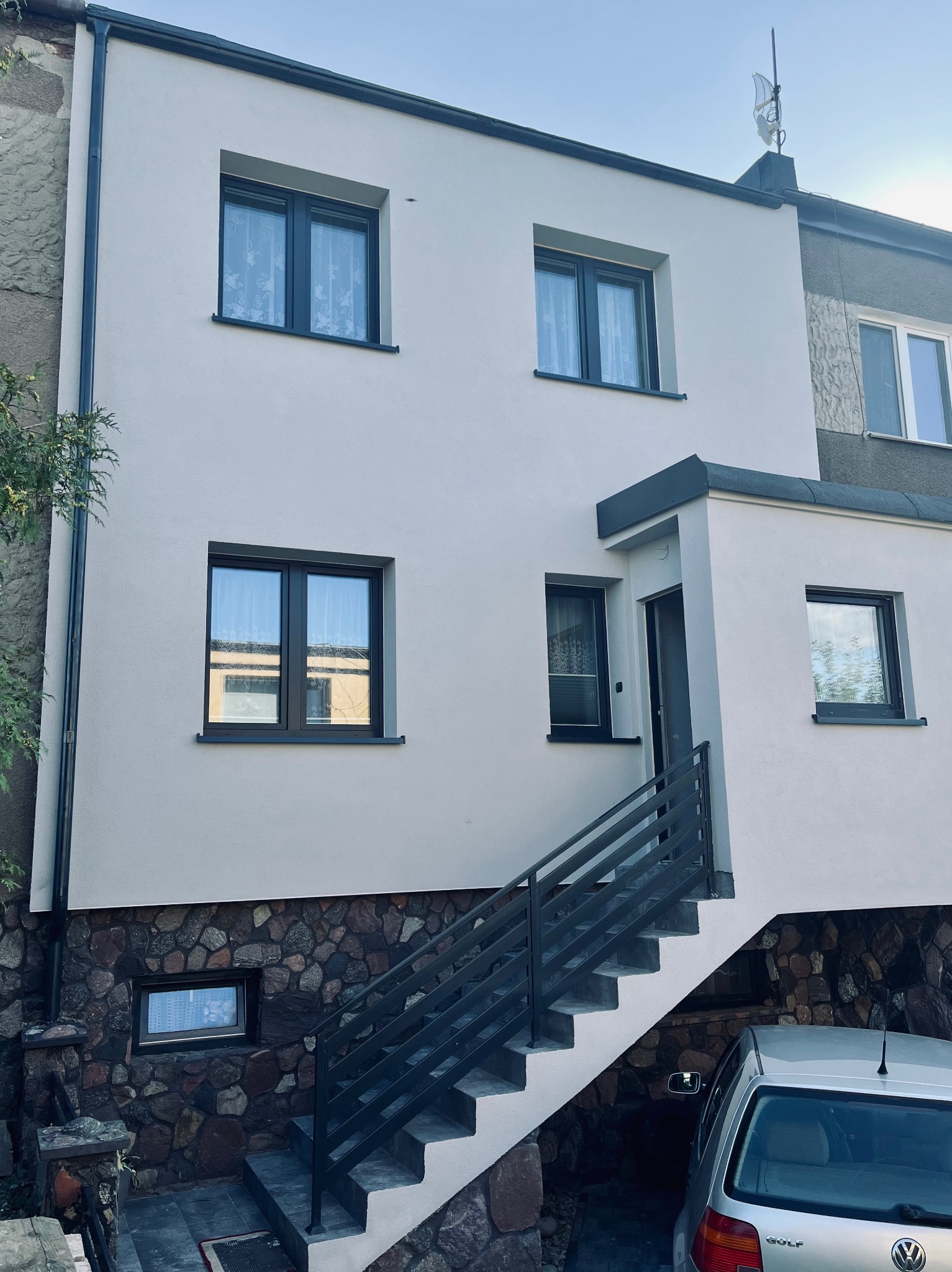 Odnowiona elewacja domu z nowymi oknami i kamienną podmurówką, schody wejściowe z metalową barierką. Widoczny samochód osobowy. Antena na dachu.