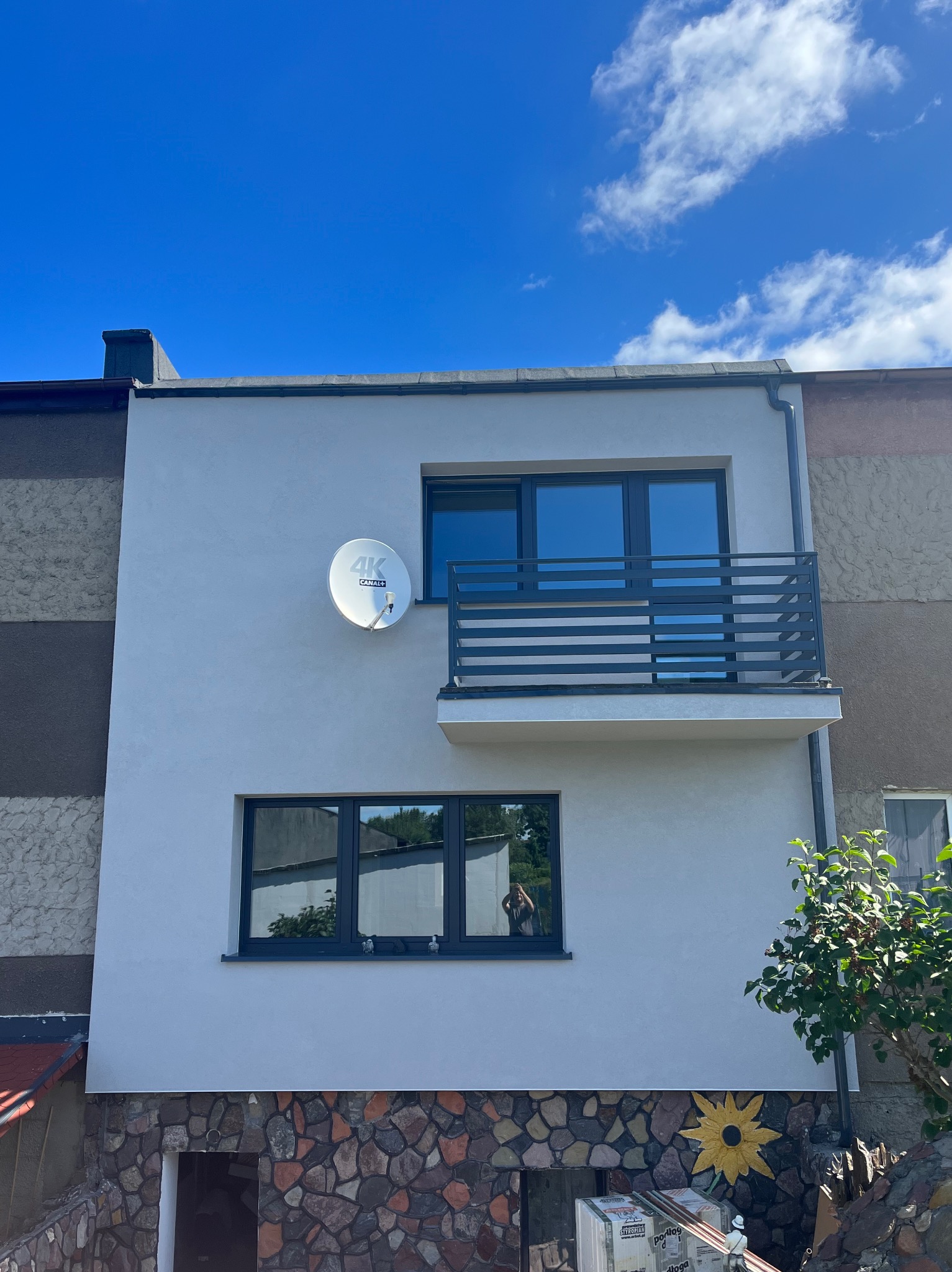 Dwukondygnacyjny dom z balkonem i anteną satelitarną na elewacji, widok z dołu na tle błękitnego nieba z chmurami. Część budynku wykończona kamieniem.