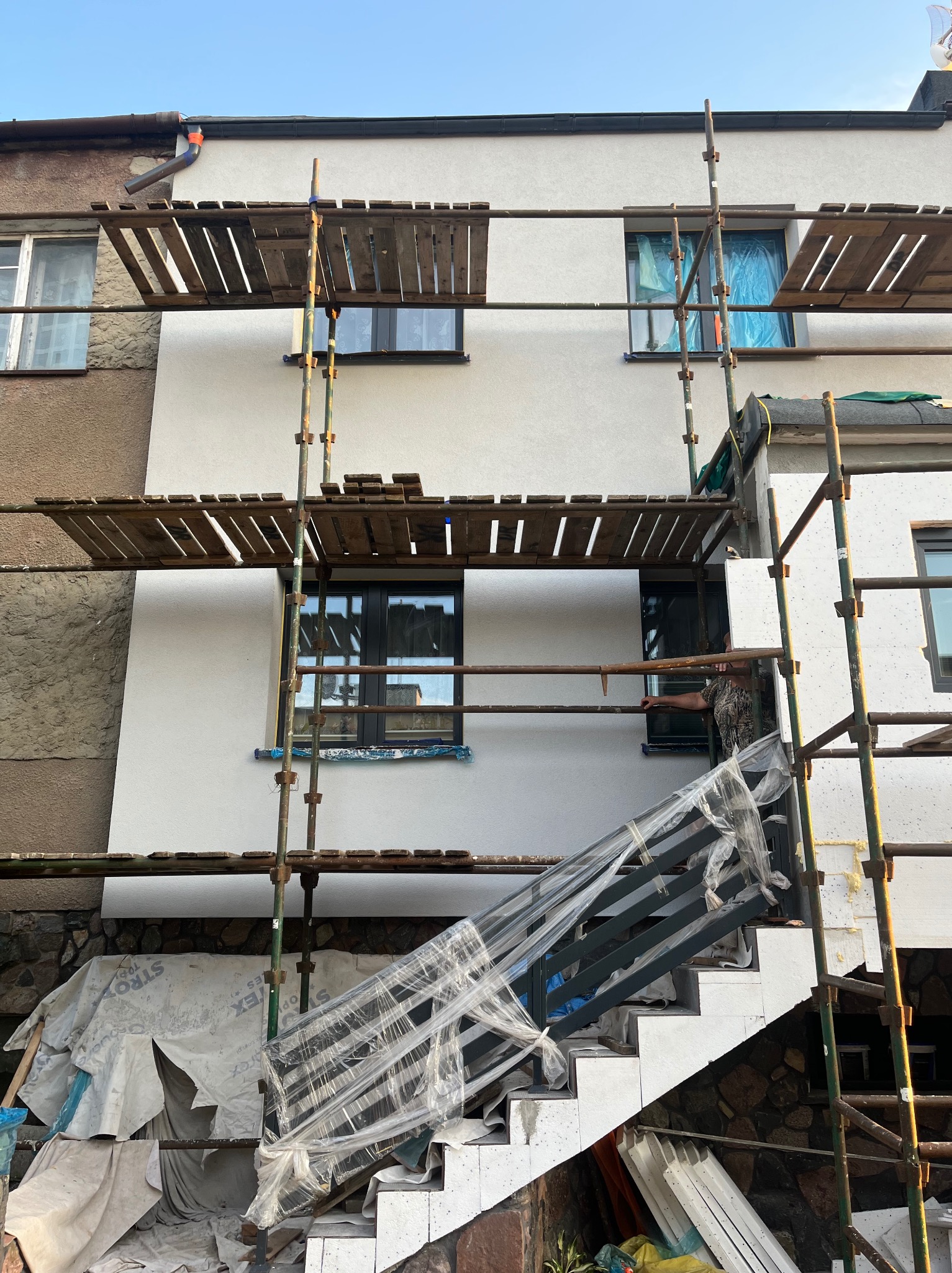 Ocieplanie elewacji domu z rusztowaniem i nowymi schodami wejściowymi. Prace wykończeniowe, styropian na ścianach, okna zabezpieczone folią.