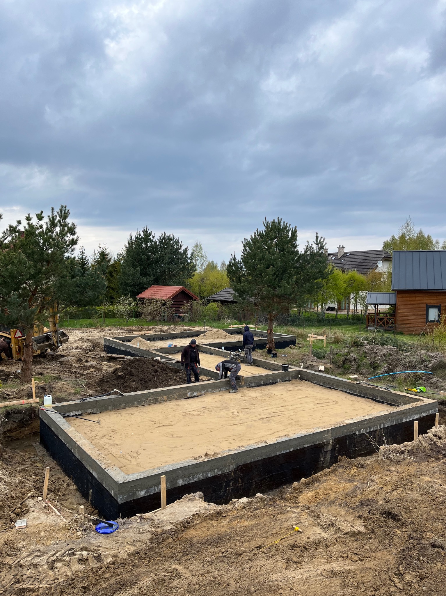 Fundamenty pod budowę domu szkieletowego, widoczni pracownicy. Zabezpieczone ściany fundamentowe, zasypane piaskiem. W tle zabudowa jednorodzinna i pochmurne niebo.
