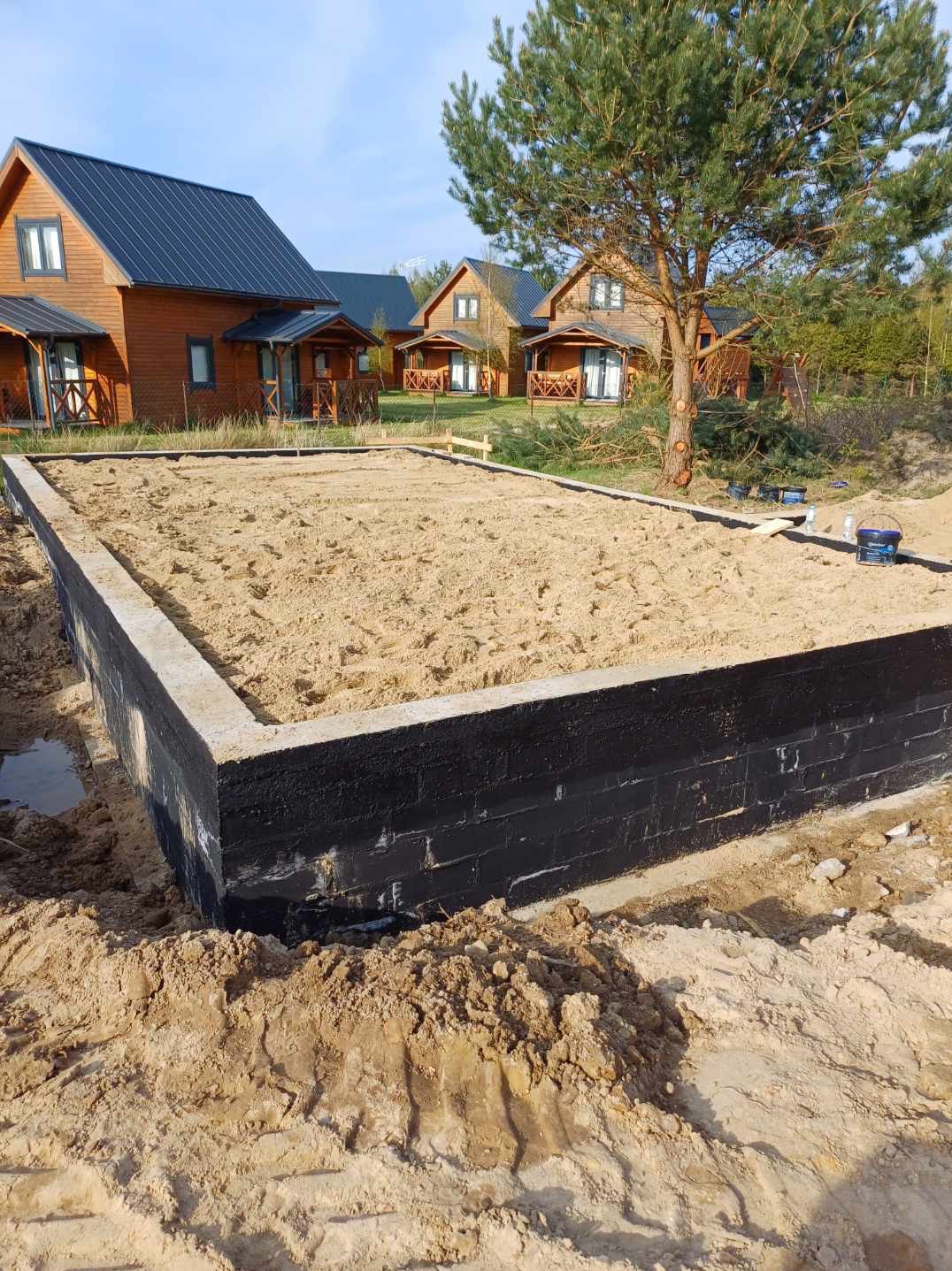 Fundament pod dom szkieletowy z czarną izolacją, wypełniony piaskiem, na tle drewnianych domków letniskowych i drzewa. Widok z dołu.