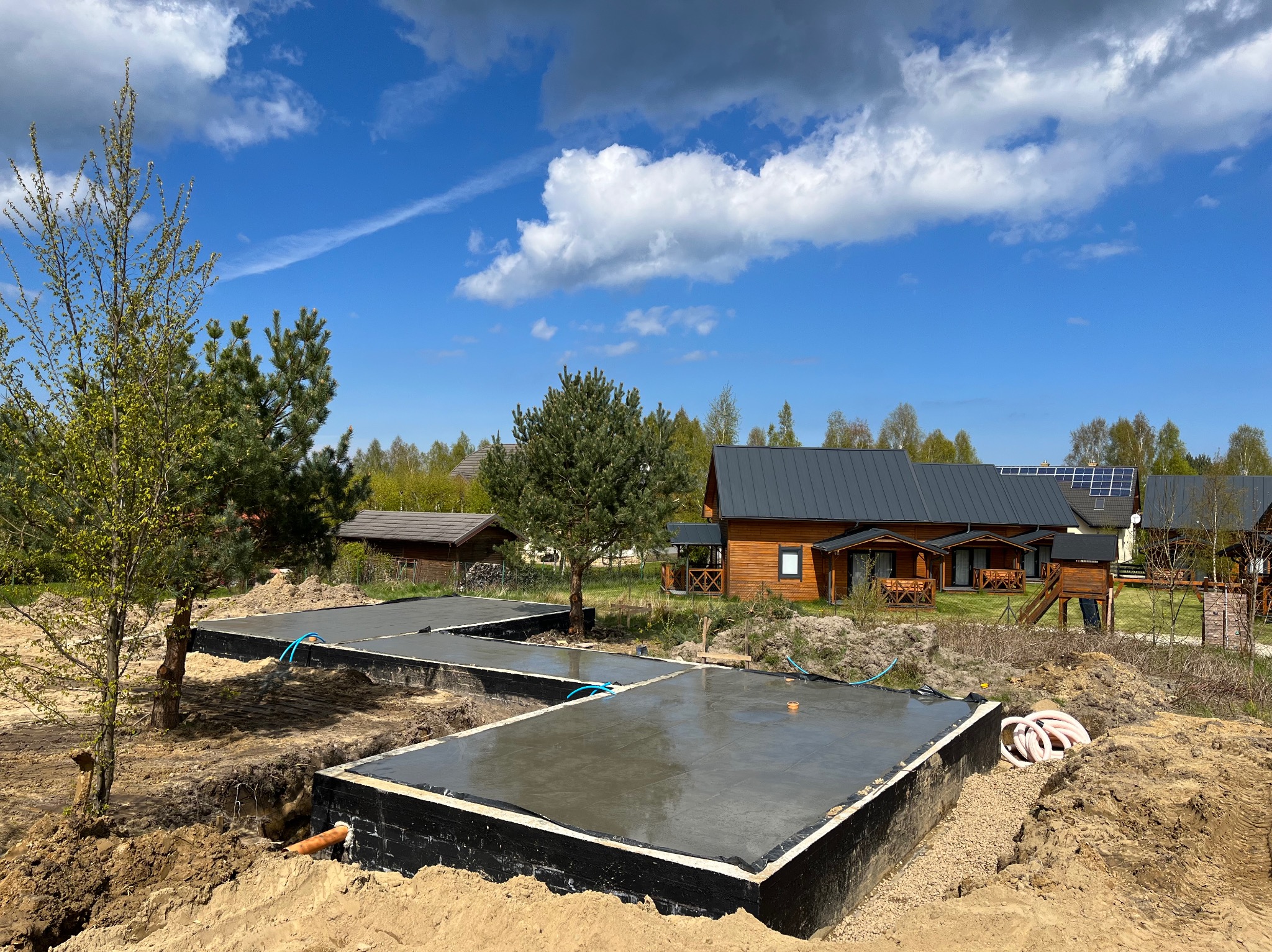 Fundamenty pod dom szkieletowy w Lubichowie, świeżo wylany beton, czarne szalunki, niebieskie rury instalacyjne. W tle domki z ciemnego drewna i błękitne niebo.