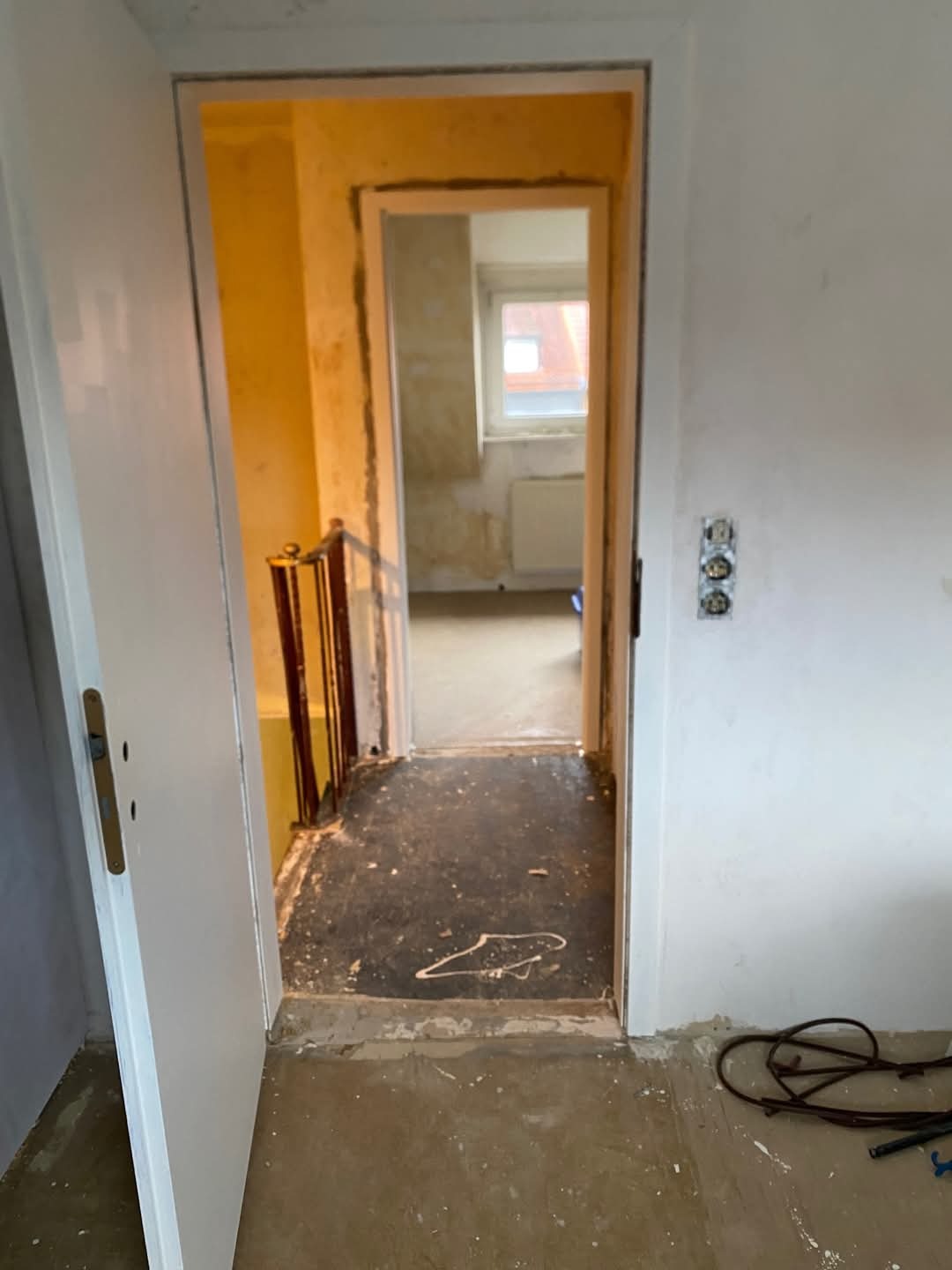 Wnętrze w trakcie remontu: widok przez otwarte drzwi na schody i dalsze pomieszczenie z oknem. Ściany w stanie surowym, widoczne ślady po pracach wykończeniowych, przewody na podłodze.