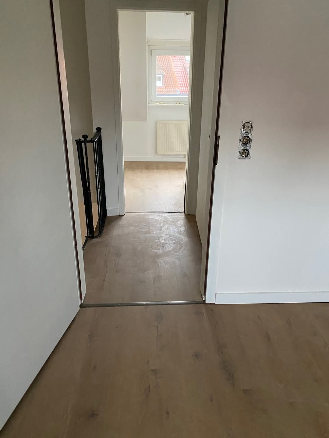 Wnętrze mieszkania po remoncie: drewniana podłoga, białe ściany, widok na pokój z oknem i grzejnikiem. Widoczne gniazdka elektryczne i fragment balustrady.