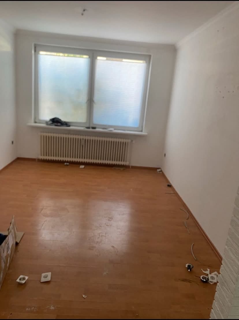 Pokój z drewnianą podłogą przed remontem, widoczne gniazdka elektryczne, grzejnik pod oknem z żaluzjami, ślady po naprawach na ścianach.