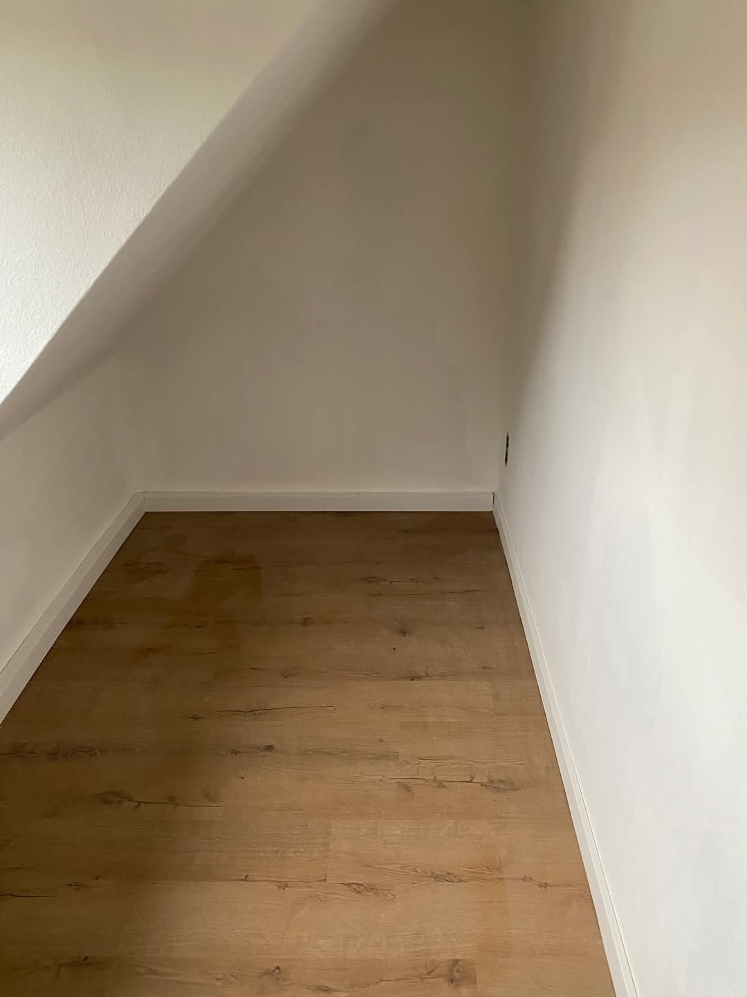 Poddasze z nową podłogą z paneli w odcieniach drewna i świeżo pomalowanymi na biało ścianami. Widoczne skosy i listwy przypodłogowe.
