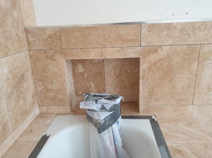 Beżowe płytki imitujące marmur, ułożone na ścianie nad wanną, tworzą wnękę. Częściowo zakryta wanna z widocznym profilem wykończeniowym.