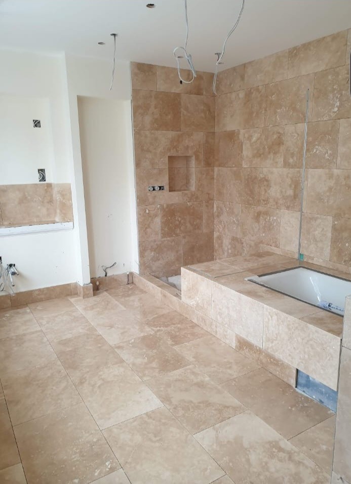 Wykończona łazienka z beżowymi płytkami trawertynowymi na podłodze i ścianach, widoczna wanna w zabudowie i instalacja elektryczna.