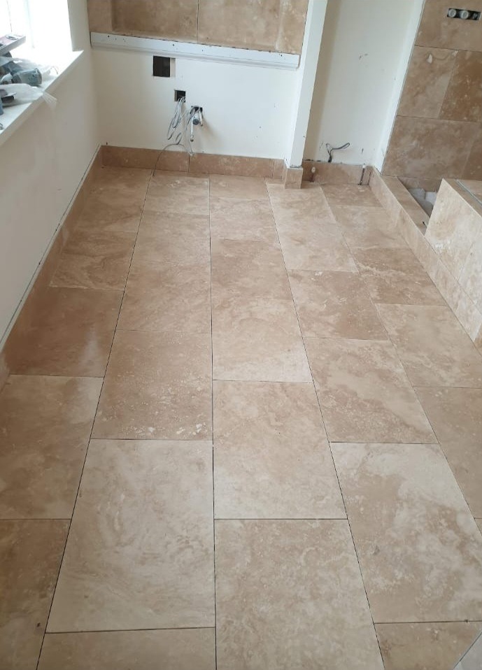 Ułożona, beżowa podłoga z płytek w trakcie prac wykończeniowych. Widoczne fragmenty ścian z instalacją elektryczną i sanitarną.