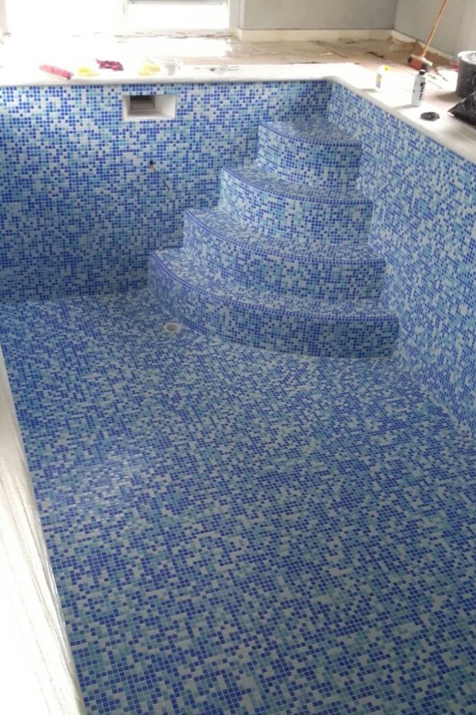 Błękitna mozaika w basenie z wyraźnymi schodami. W tle widoczne okno i akcesoria. Całość sprawia wrażenie świeżo wykończonego wnętrza.