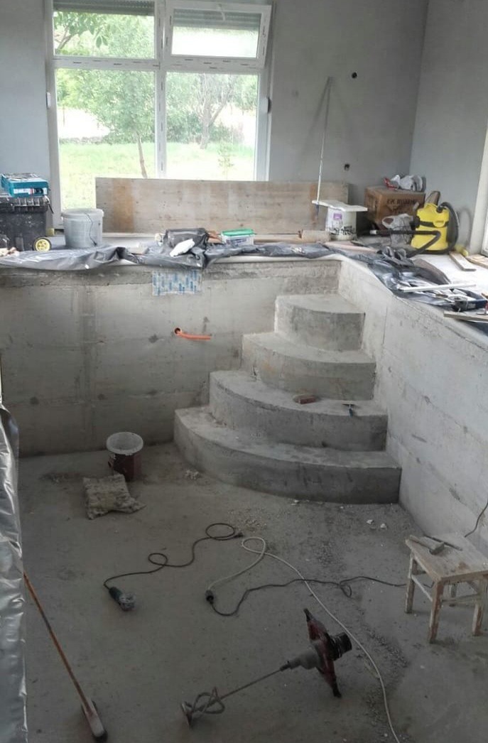 Surowy stan betonowego basenu wewnątrz budynku z oknem, widoczne schody i narzędzia budowlane. Etap budowy przed wykończeniem.