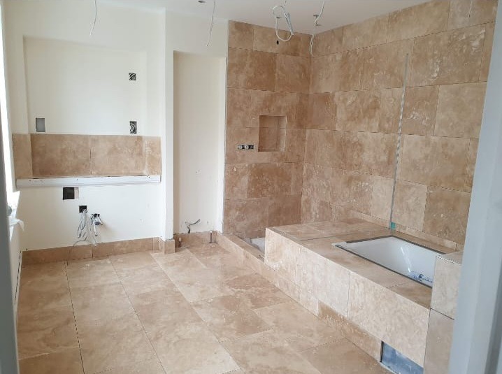 Beżowe płytki ścienne i podłogowe w łazience w trakcie wykończenia. Widoczne przygotowane instalacje i wanna. Oświetlenie sufitowe w postaci zwisających kabli.