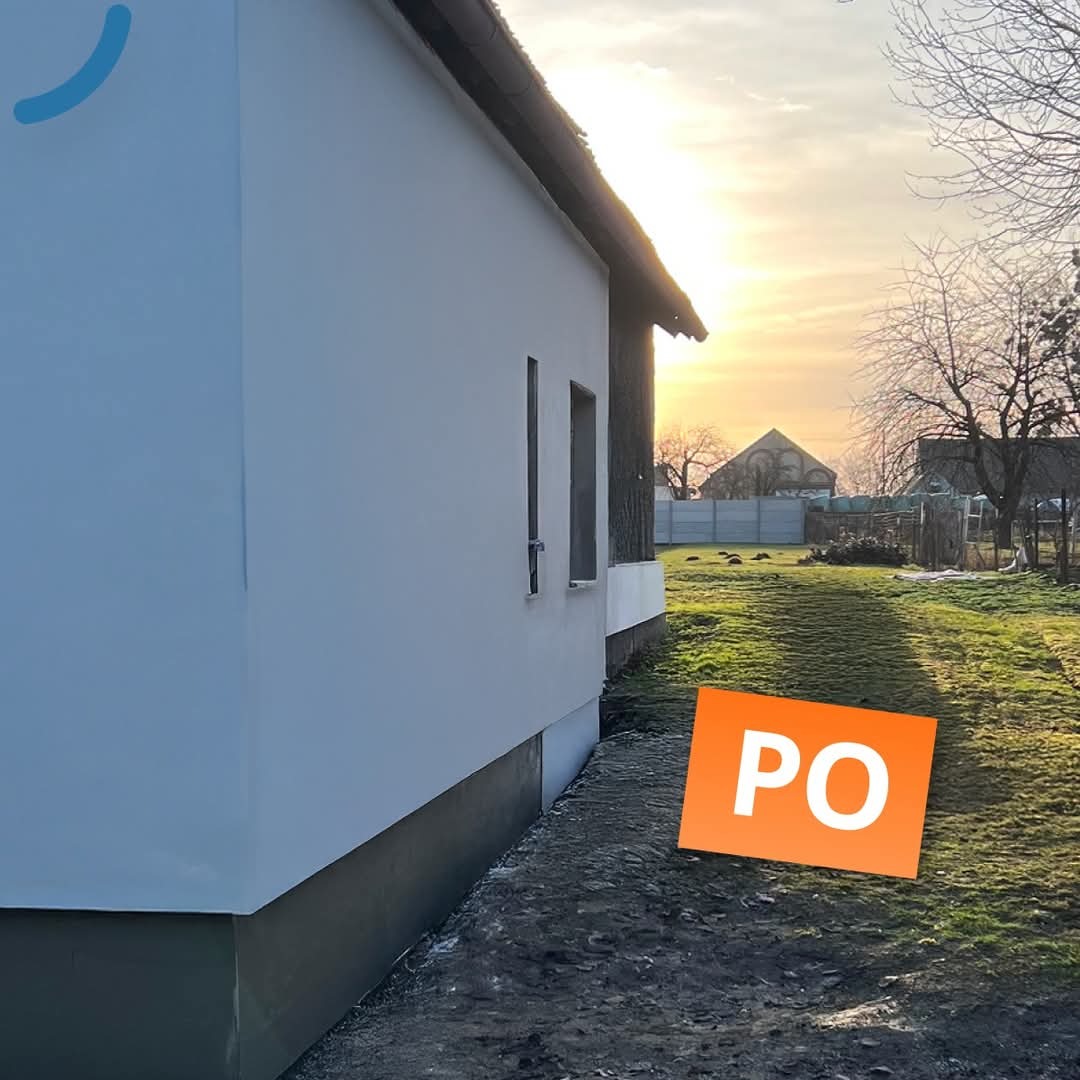 Elewacja domu po remoncie, widok z boku, czysta, biała ściana, oświetlona zachodzącym słońcem, zieleń trawy w tle, pomarańczowa nakładka 'PO'.