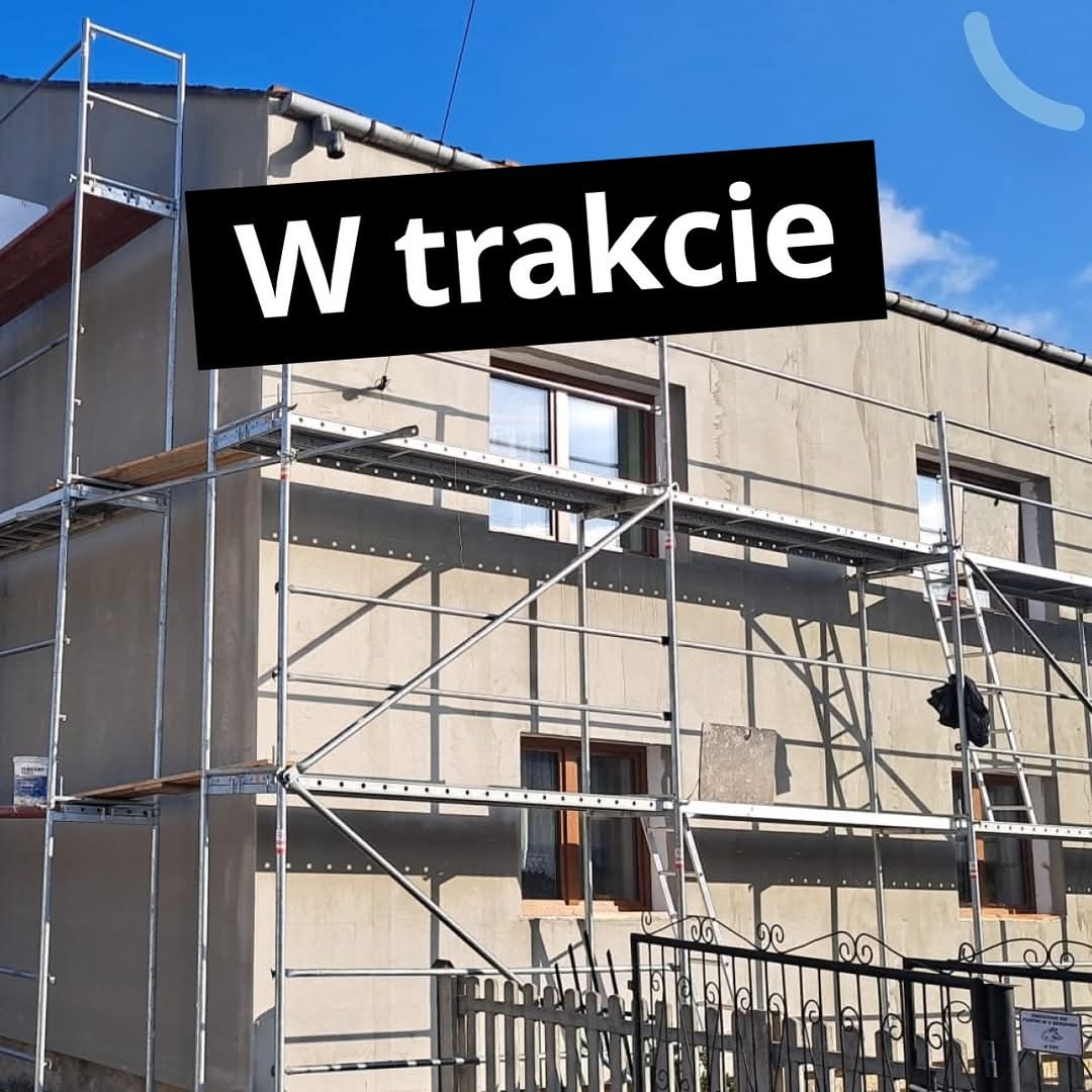 Dom w trakcie prac elewacyjnych, otoczony rusztowaniem, z widocznym napisem 'W trakcie' nałożonym na zdjęcie. Prace postępują, widoczne okna i ogrodzenie.
