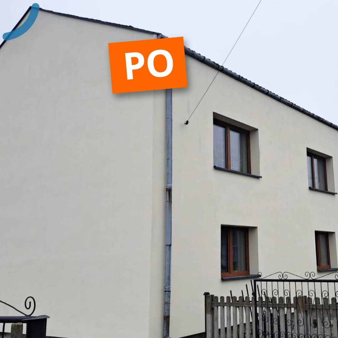 Elewacja budynku po remoncie, z nową, jasną farbą. Widoczne okna z brązowymi ramami i ozdobne ogrodzenie. Na elewacji widoczny napis 'PO' na pomarańczowym tle.