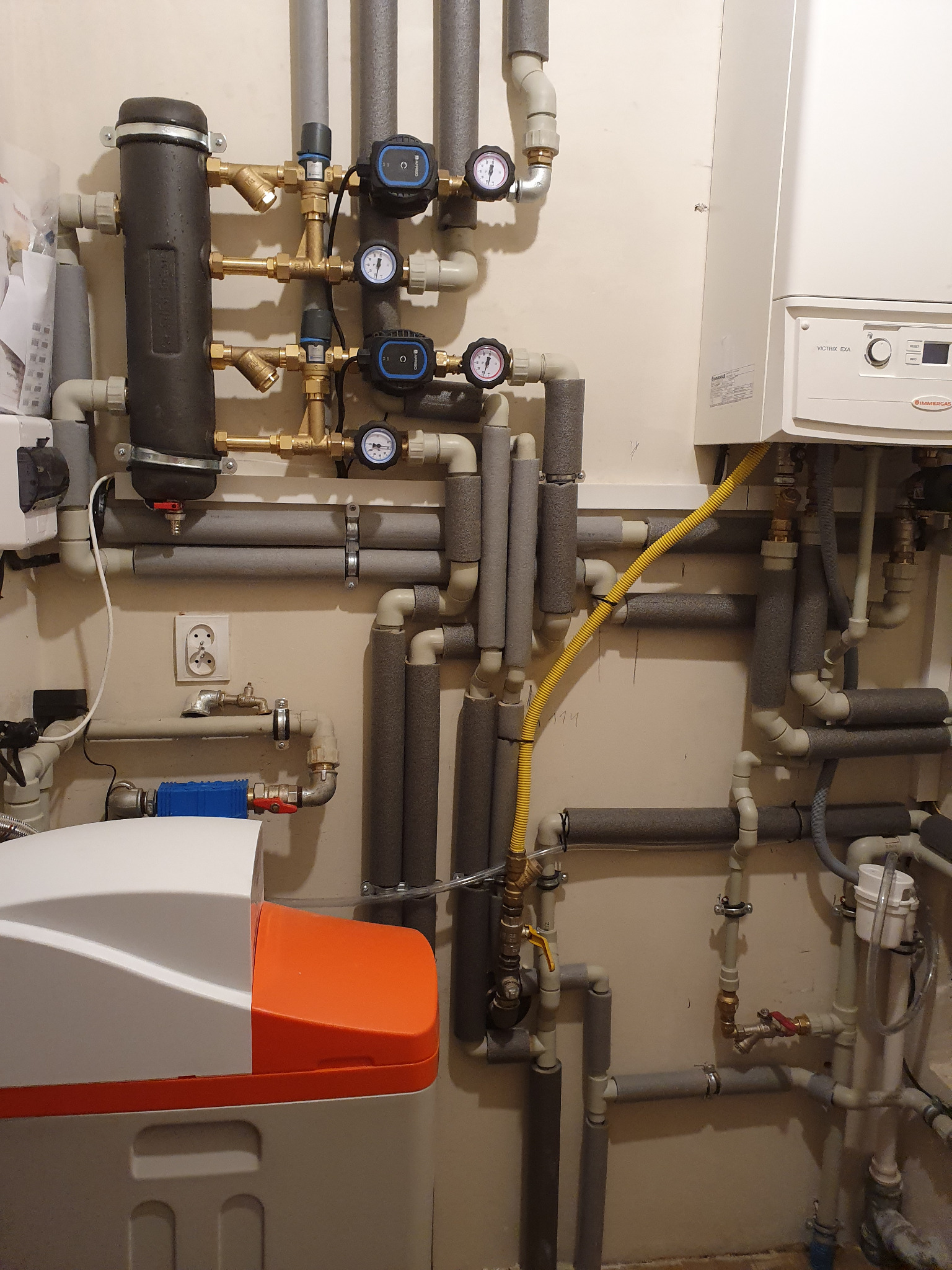 Złożona instalacja grzewcza z piecem gazowym Immergas, rurami zaizolowanymi szarą pianką i filtrem wody z pomarańczową pokrywą. Żółty przewód gazowy podłącza piec.