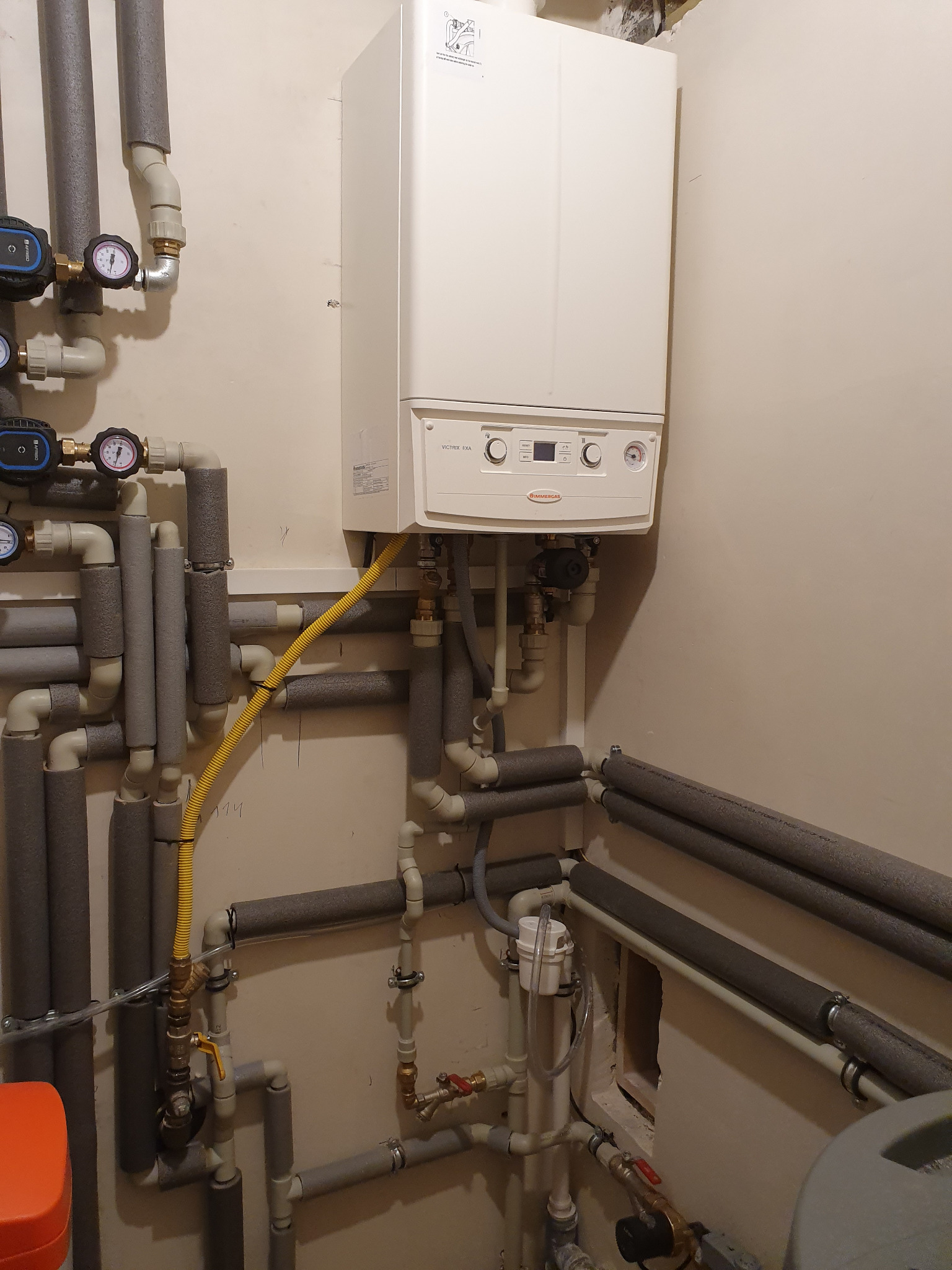 Biały piec gazowy Immergas z podłączonymi rurami w izolacji, żółty przewód gazowy, manometry i zawory. Instalacja w jasnym pomieszczeniu.