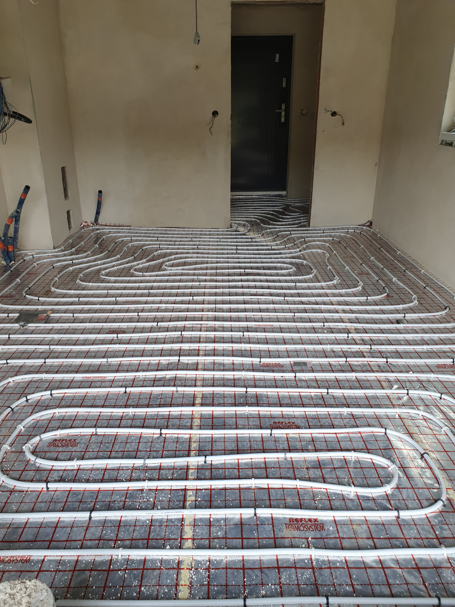 Białe rury ogrzewania podłogowego ułożone na czarnej macie z czerwoną siatką, widok na próg drzwi wejściowych. Etap instalacji w surowym wnętrzu.