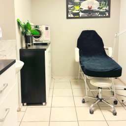 Gabinet Kosmetyki Laserowej i Stylizacji Paznokci Dorota Musidlak - Jasne wnętrze gabinetu kosmetycznego z fotelem zabiegowym, sterylizatorem i rośliną w doniczce. Na ścianie obraz z motywem kwiatowym.