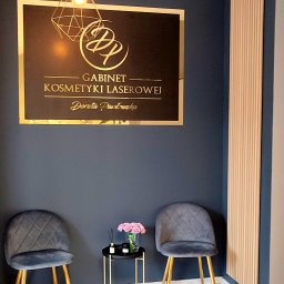 Gabinet Kosmetyki Laserowej i Stylizacji Paznokci Dorota Musidlak - Elegancki gabinet kosmetyki laserowej z logo, dwoma krzesłami i stolikiem z kwiatami na tle ściany w kolorze granatowym. Oświetlenie punktowe.