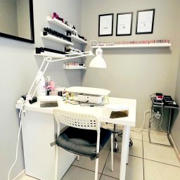 Gabinet Kosmetyki Laserowej i Stylizacji Paznokci Dorota Musidlak - Stanowisko do manicure w salonie kosmetycznym: białe biurko z lampą, pochłaniacz pyłu, lakiery na półkach i certyfikaty na ścianie w minimalistycznym wnętrzu.