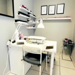 Stanowisko do manicure w salonie kosmetycznym: białe biurko z lampą, pochłaniacz pyłu, lakiery na półkach i certyfikaty na ścianie w minimalistycznym wnętrzu.