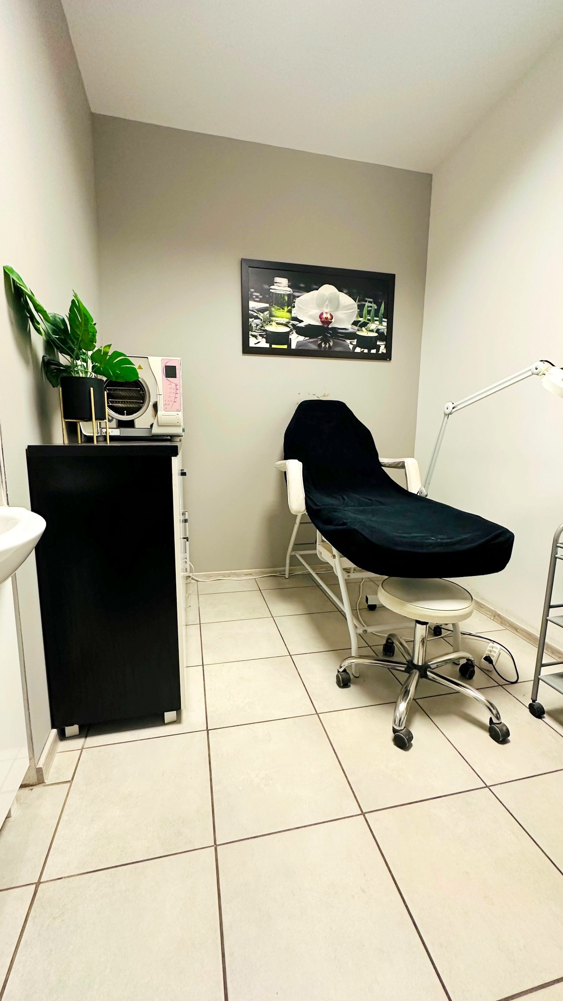 Nowoczesny gabinet kosmetyczny: czarne obicie fotela, sterylizator, roślina doniczkowa i grafika na ścianie. Jasne płytki podłogowe, lampa kosmetyczna.