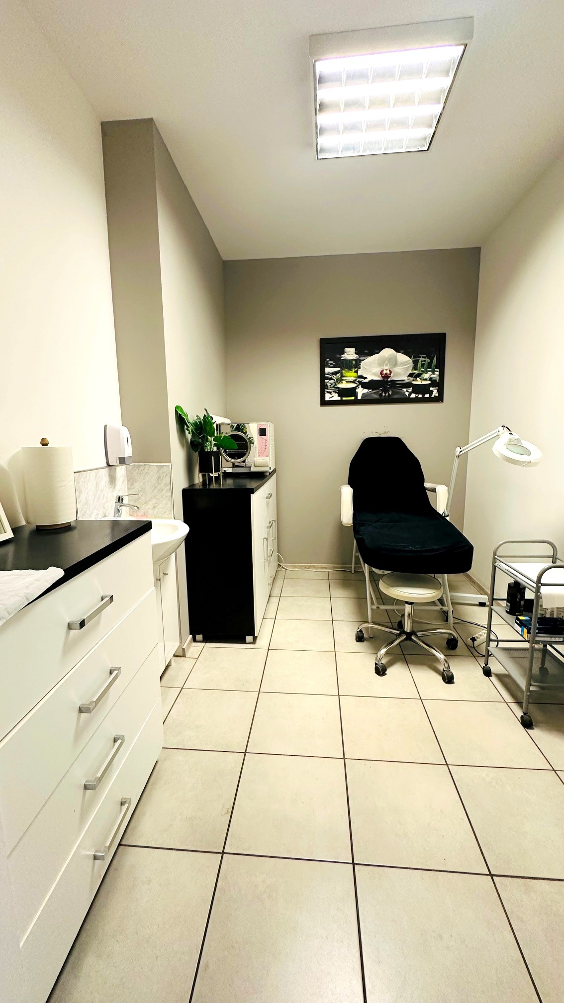 Jasne wnętrze gabinetu kosmetycznego z fotelem zabiegowym, szafkami i lampą powiększającą. Na ścianie wisi obraz z motywem kwiatowym.