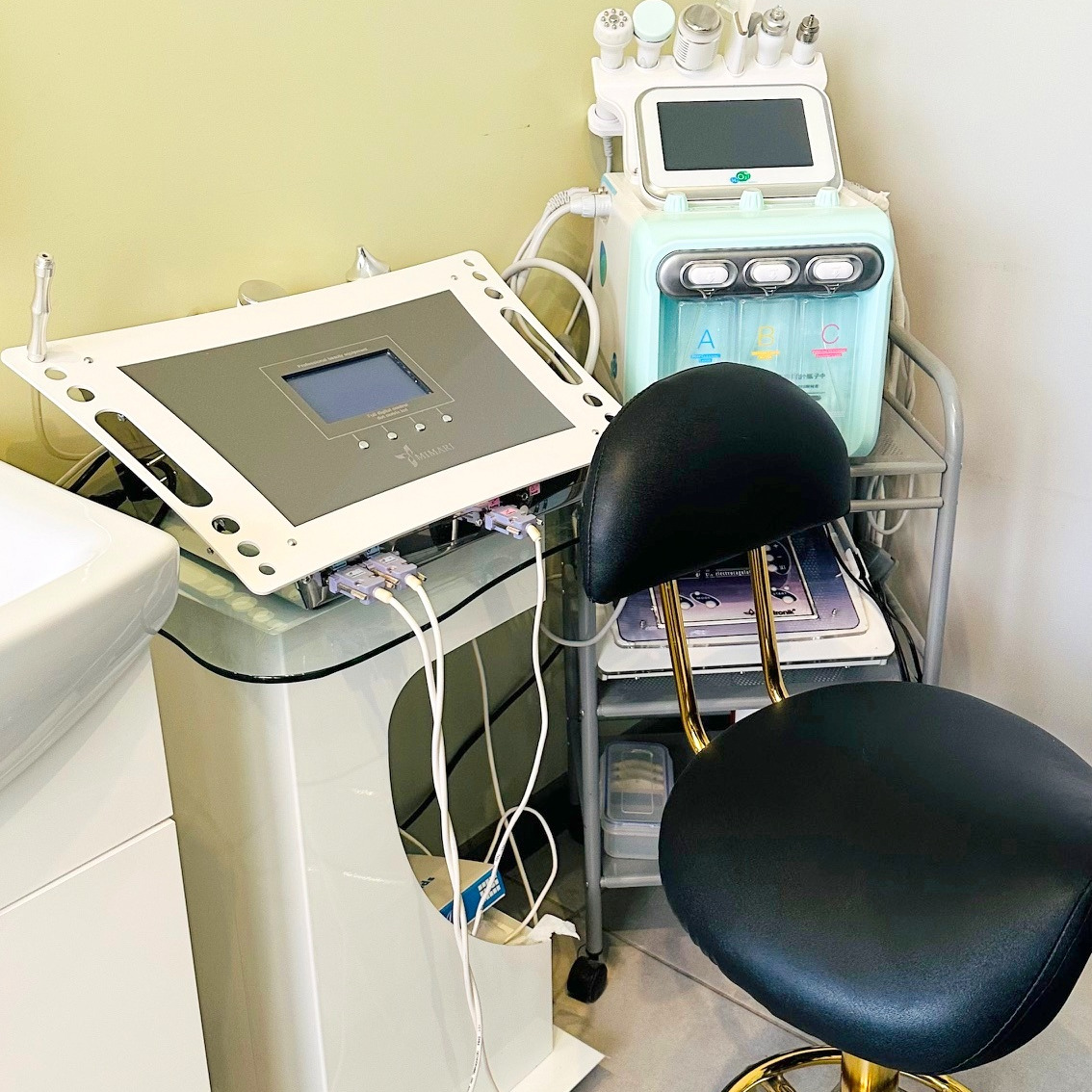 Wyposażenie gabinetu kosmetycznego: dwa urządzenia z ekranami, krzesło z czarnym siedziskiem i złotymi nogami, ustawione w rogu pomieszczenia.