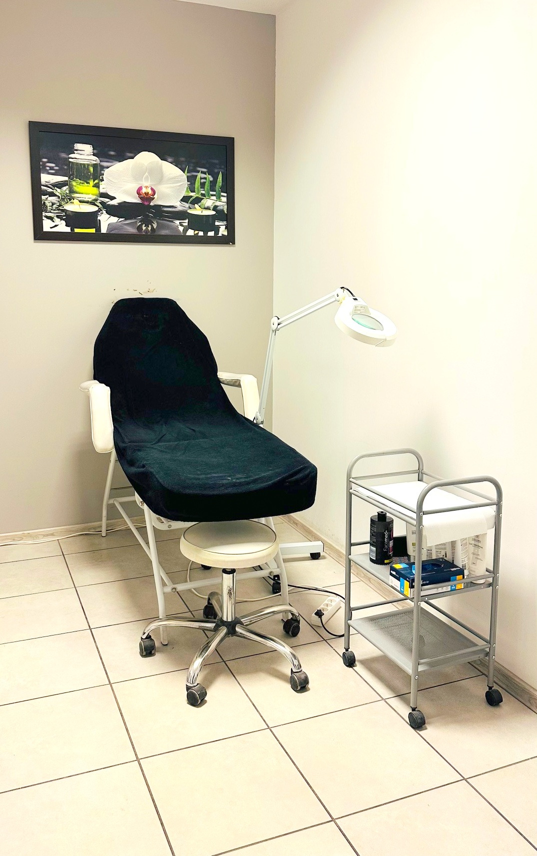 Kącik kosmetyczny: fotel z czarnym pokrowcem, lampa powiększająca i wózek z kosmetykami. Na ścianie obraz z motywem orchidei i świec. Jasne płytki na podłodze.