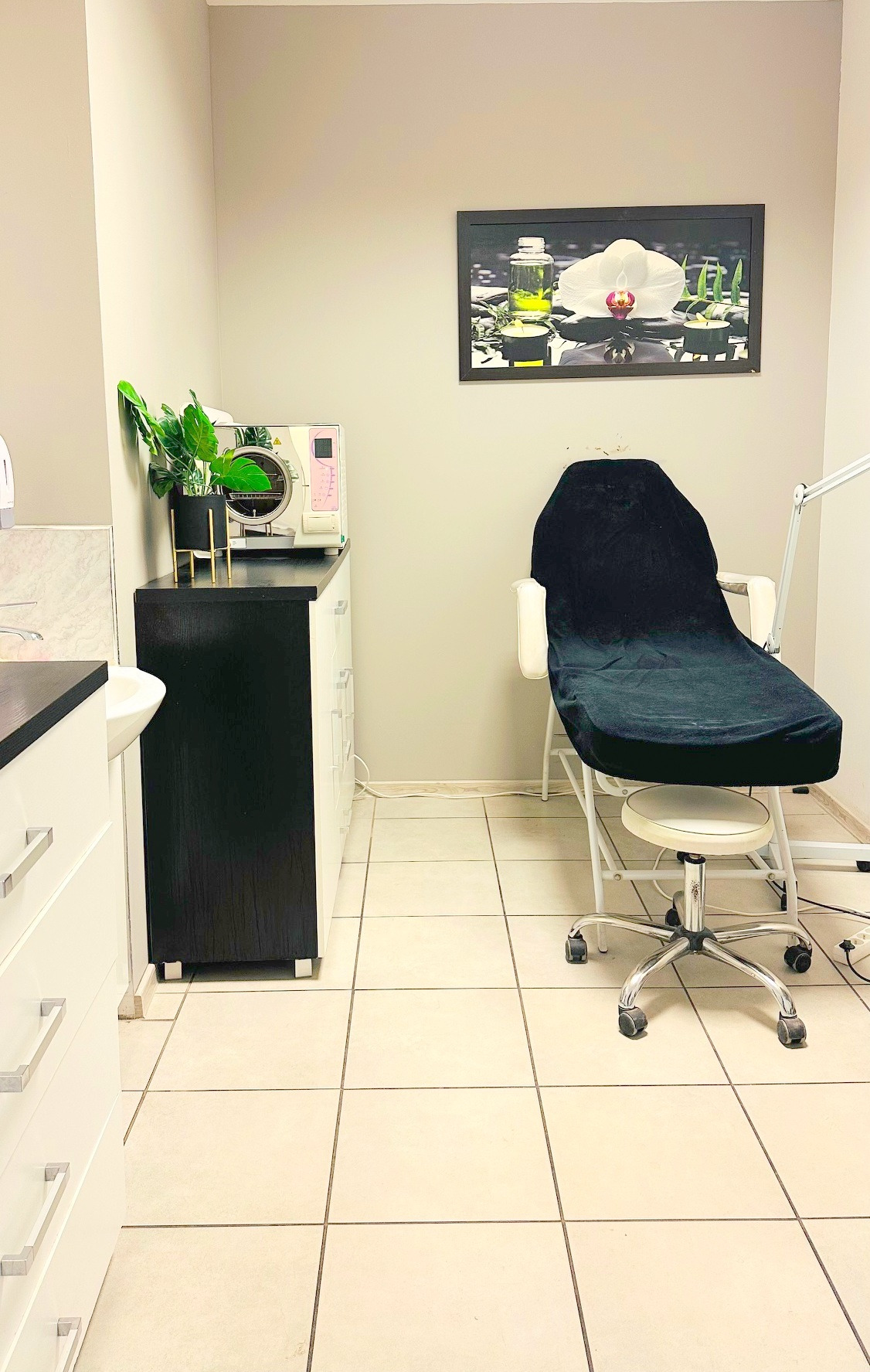Jasne wnętrze gabinetu kosmetycznego z fotelem zabiegowym, sterylizatorem i rośliną w doniczce. Na ścianie obraz z motywem kwiatowym.