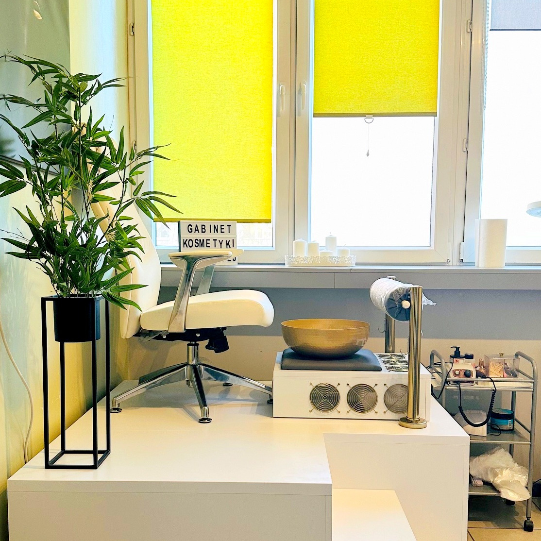 Wnętrze gabinetu kosmetycznego z nowoczesnym stanowiskiem pracy, rośliną w czarnej donicy i oknem z żółtymi roletami w tle.