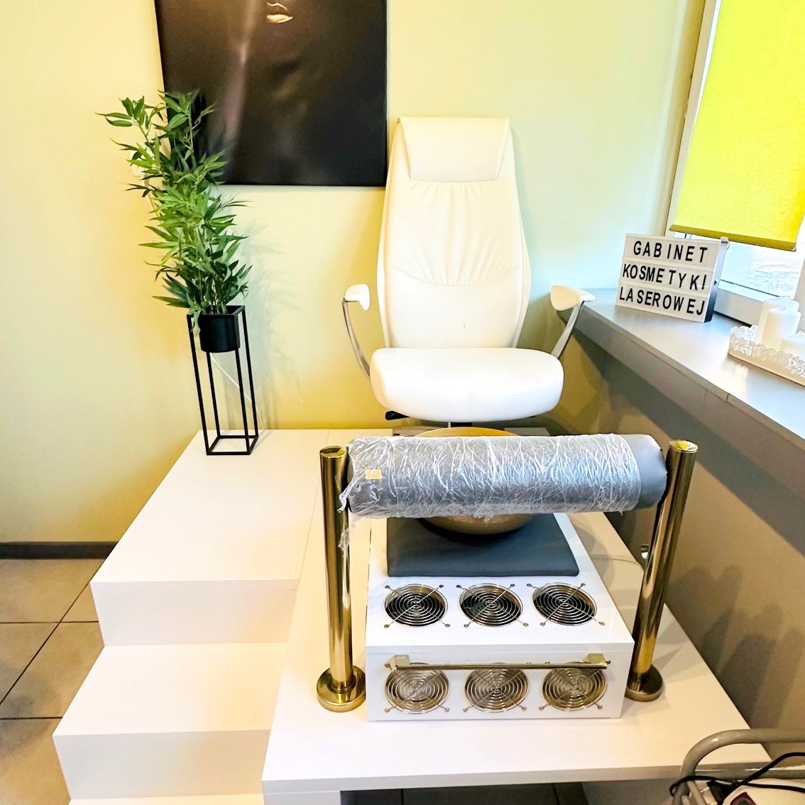 Wnętrze gabinetu kosmetyki laserowej: biały fotel, urządzenie z wentylatorami i złotymi detalami, roślina w czarnej doniczce, dekoracje okienne.
