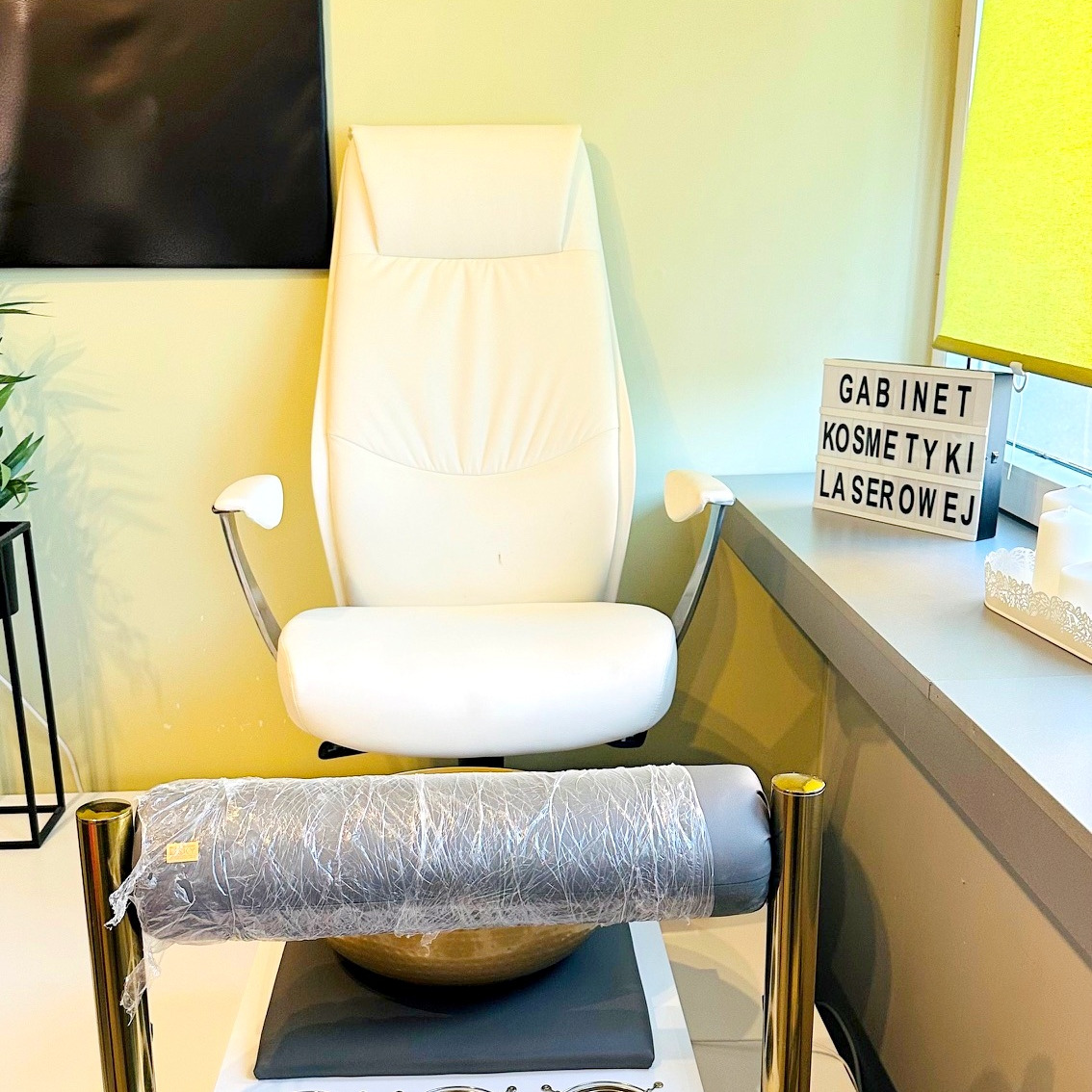 Jasny fotel do pedicure w gabinecie kosmetycznym, widoczna wanienka do stóp i dekoracyjny napis 'GABINET KOSMETYKI LASEROWEJ'.
