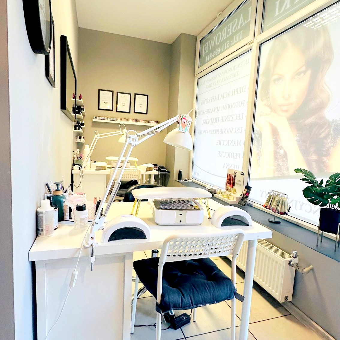 Jasne wnętrze salonu z białymi stanowiskami do manicure, lampami UV i wzornikami lakierów przy oknie z reklamą. Minimalistyczny wystrój, czyste linie.