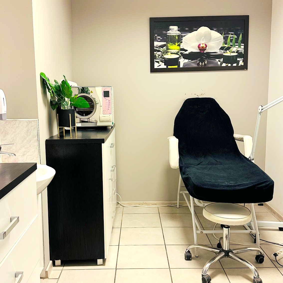 Wnętrze gabinetu kosmetycznego z fotelem zabiegowym pod czarnym pokrowcem, sterylizatorem, umywalką i dekoracją kwiatową na ścianie.